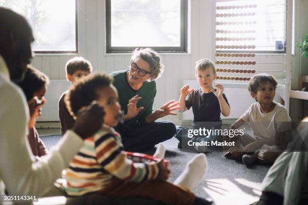senior female teacher playing with group of preschool children while sitting in classroom at kindergarten - peuterschool gebouw stockfoto's en -beelden