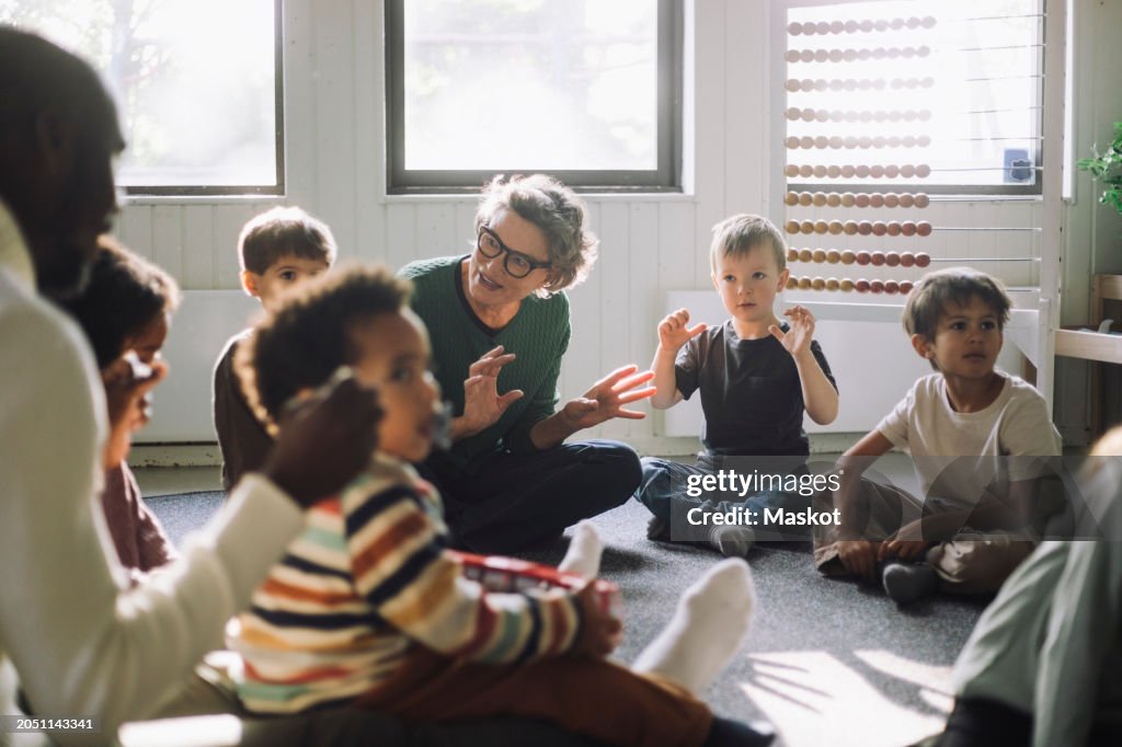 Senior female teacher playing with group of preschool children while sitting in classroom at kindergarten