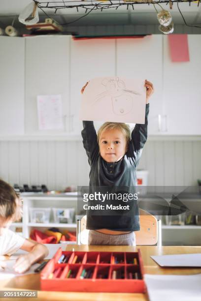 portrait of girl showing paper with drawing while standing near bench in classroom - animal representation stock pictures, royalty-free photos & images