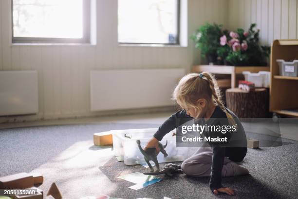 side view of girl playing with artificial dinosaurs while sitting on carpet in classroom at kindergarten - animal representation stock pictures, royalty-free photos & images