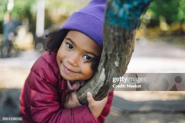 portrait of smiling girl wearing knit hat and embracing tree branch at park - stockholm park stock pictures, royalty-free photos & images