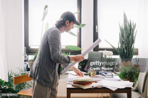 side view of male architect holding rolled blueprint while standing at desk in home office - book icon stock pictures, royalty-free photos & images