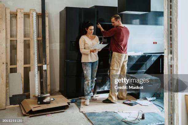 couple discussing over document while fixing cabinet together in kitchen during home renovation - renovierung themengebiet stock-fotos und bilder