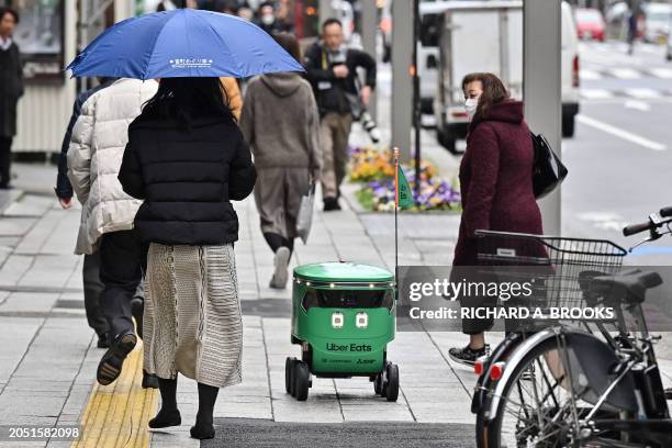 Pedestrians walk alongside an unmanned robot as it navigates down a street during a demonstration of a robot delivery service by Uber Eats Japan,...