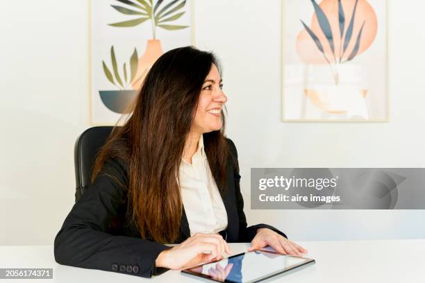 smiling woman sitting in front of her desk with her hands on her notebook looking at the camera behind her is a white wall with beautiful decorative posters. - chairperson stock pictures, royalty-free photos & images