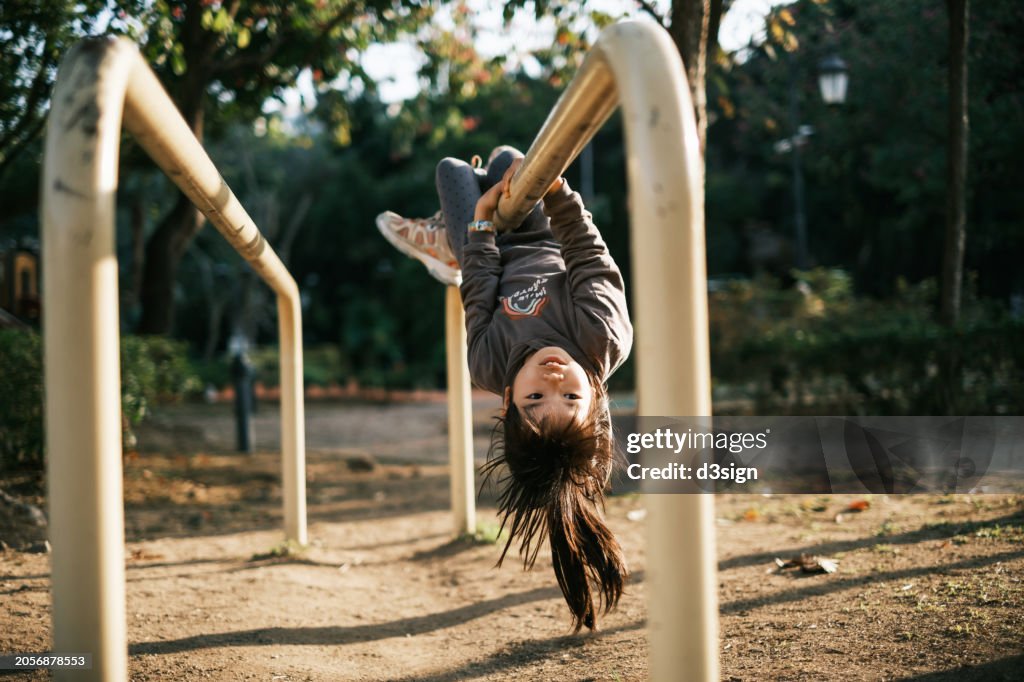 Playful little Asian girl hanging upside down on a parallel bar, having fun at the outdoor playground on a sunny day. Self-learning. Outdoor fun activity concept
