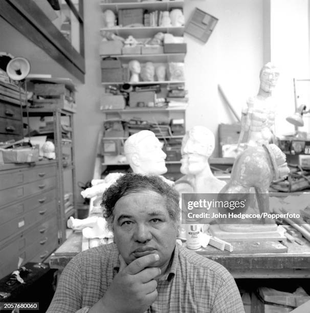 Scottish artist and sculptor Eduardo Paolozzi posed in front of various cast heads and works in progress in his studio workplace in Chelsea, London...