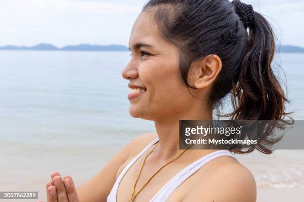 portrait of thai woman on the beach practicing yoga, she holds the namaste prayer pose on her heart center, she smiles - prayer pose greeting stock-fotos und bilder