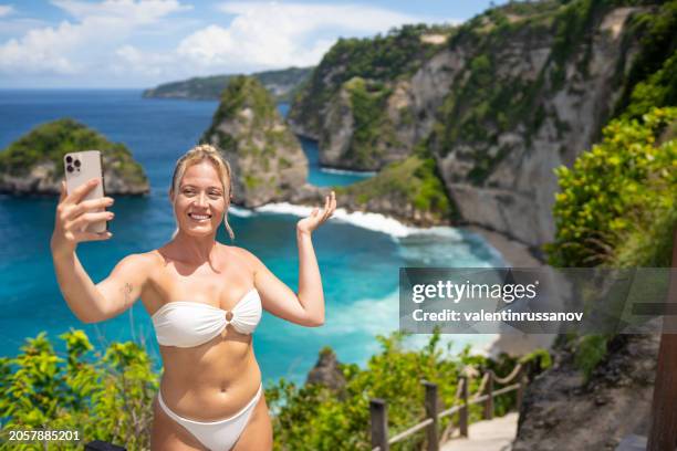 young female tourist in swimsuit taking selfie on kelingking beach on nusa penida island, bali, indonesia - indonesien stock-fotos und bilder