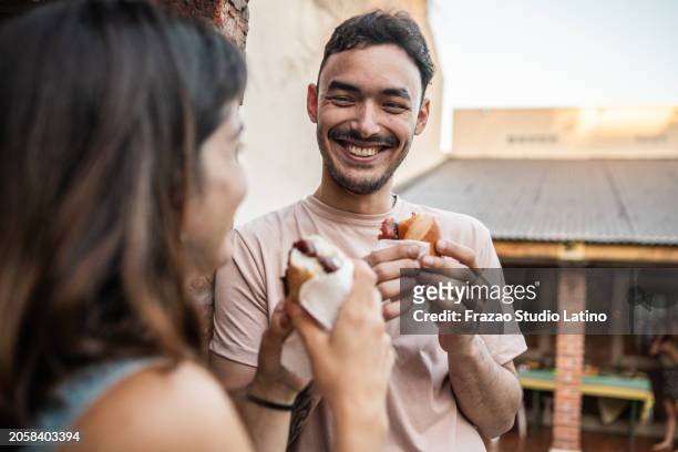 young man talking and eating choripán with friend in the backyard at home - barbecue sandwich stock pictures, royalty-free photos & images