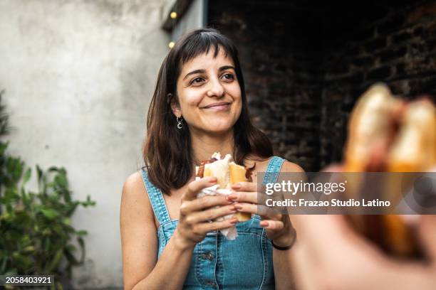 mid adult woman talking and eating choripán with friend in the backyard at home - chorizo stock pictures, royalty-free photos & images