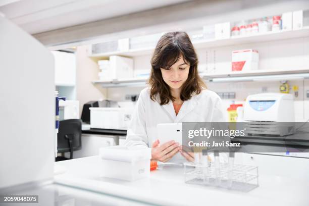 young woman scientist in a lab coat examining information on a tablet while working with test tubes in the laboratory. - modern laboratory stock pictures, royalty-free photos & images