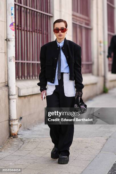 Guest wears red sunglasses, a blue shirt, a tie, a black jacket , black pants , leather shoes, outside Marine Serre, during the Womenswear...