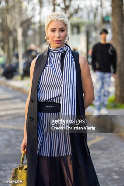 Guest wears blue white striped dress, black sleeveless coat, transparent belted skirt outside Sacai during the Womenswear Fall/Winter 2024/2025 as...