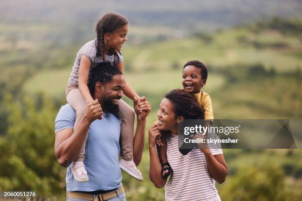 padres negros felices que llevan a sus hijos pequeños sobre hombros en la naturaleza. - llevar al hombro fotografías e imágenes de stock