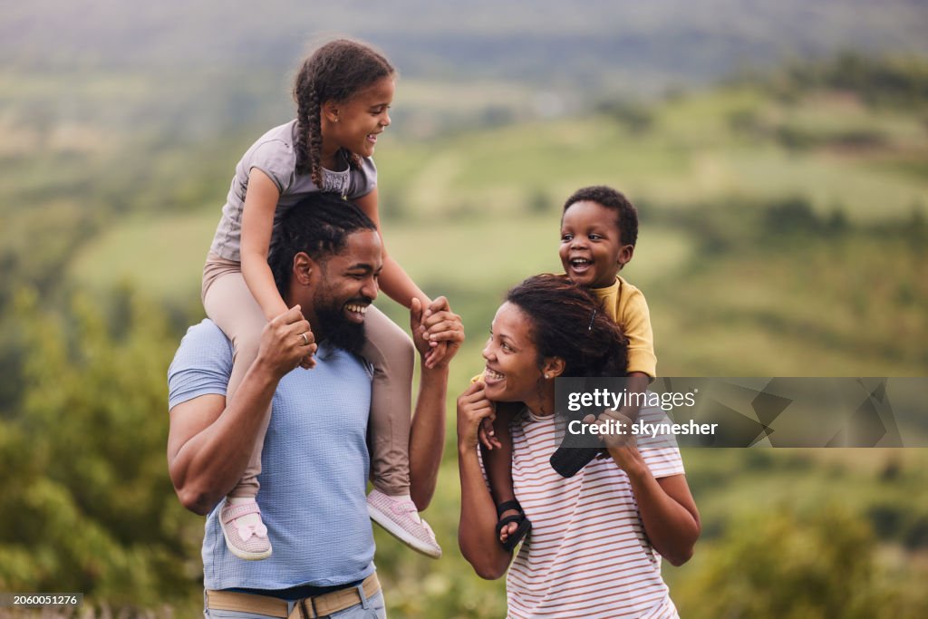 Happy black parents carrying their small kids on shoulders in nature.