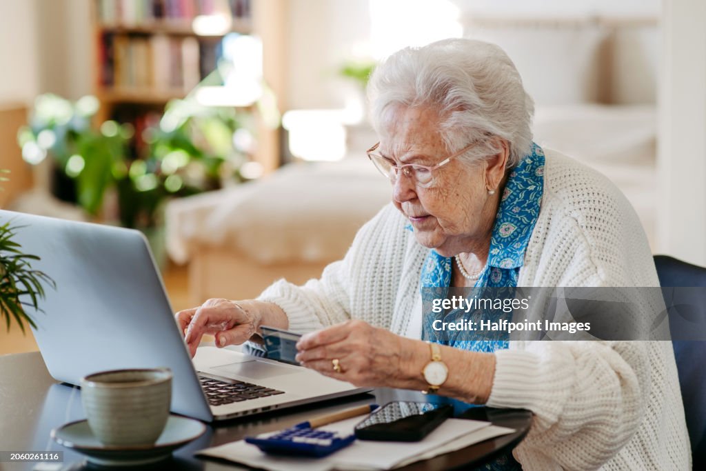 Senior woman paying with debit card online, using internet banking and payment gateway. Elderly woman working on laptop, shopping online, risk of frauds and hoax for elderly people.