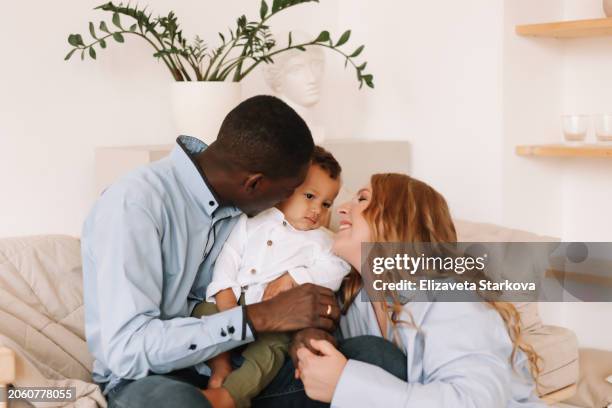 a multiracial family with one small child in casual clothes hugging, kissing and playing in a cozy room at home. family lifestyle. a diverse group of people - family and happiness and diverse imagens e fotografias de stock