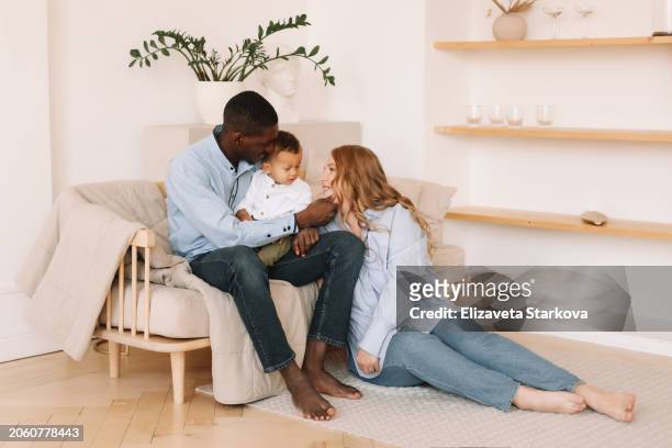 a multiracial family with one small child in casual clothes hugging, kissing and playing in a cozy room at home. family lifestyle. a diverse group of people - family and happiness and diverse imagens e fotografias de stock
