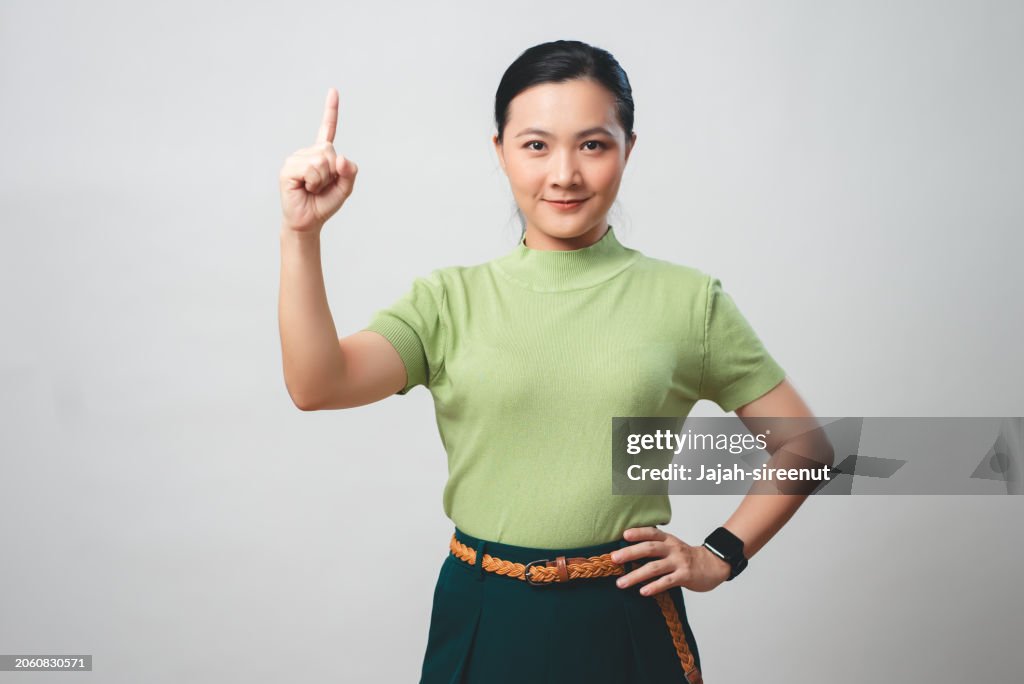Mujer asiática sonriendo con confianza señalando con el dedo hacia arriba mostrando el número uno aislado en el fondo.