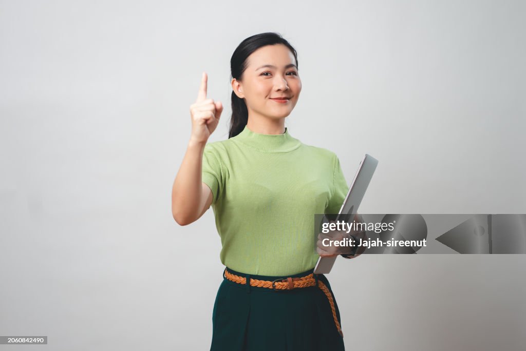 Mujer asiática sosteniendo una computadora portátil señalando con el dedo hacia arriba que muestra el número uno, aislada en el fondo.