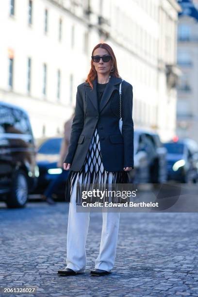 Alice Barbier wears sunglasses, a black oversized blazer jacket, a mesh fringed dress, white pants , black leather pointed shoes, outside Sacai,...