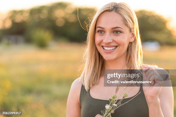 portrait of beautiful blond woman holding a grass straw, looking at the camera in front of grass field during sunset - stralende lach stockfoto's en -beelden