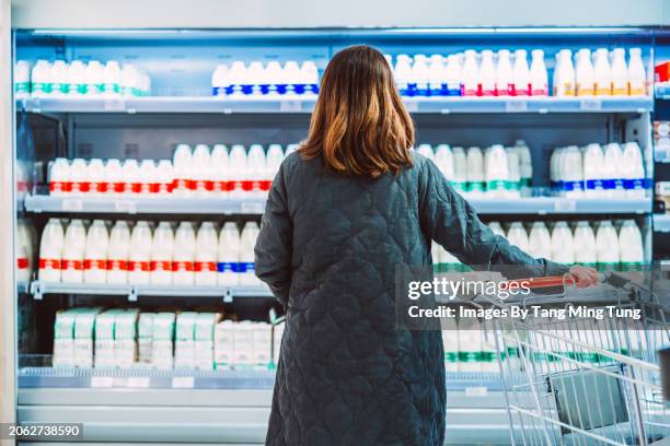rear view shot of a female customer looking and choosing fresh bottled milk while standing in front of a display fridge full of diary products in supermarket. the concept of choice. - producto lácteo fotografías e imágenes de stock