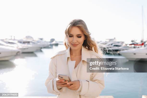 a happy smiling woman is typing a message using a mobile phone and enjoying a walk alone against the background of yachts and the sea on a travel - fashion industry stock pictures, royalty-free photos & images