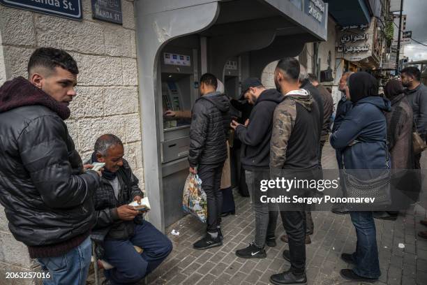 Palestinians who exchange money sit next to a line of people taking money from the ATM in Ramallah, on March 7 West Bank. The Palestinian Authority...