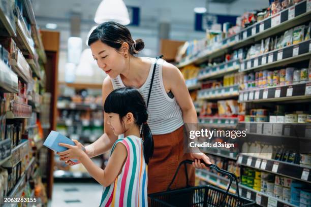 young asian mother and little daughter shopping at supermarket. they are standing along the aisle and reading the information on the package of a product, making a healthier food choice for the family. routine grocery shopping. home shopping with kid - ambientalista foto e immagini stock