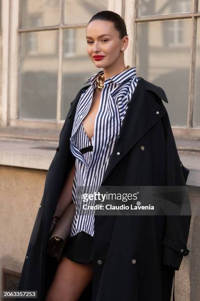 Kristina Romanova wears blue and white striped shirt, black mini skirt, black trench coat, gold necklace, outside Sacai, during the Womenswear...