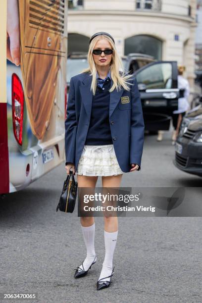 Leonie Hanne wears oversized navy blazer, white skirt, jumper, black bag, white knee high socks, black heels outside Miu Miu during the Womenswear...