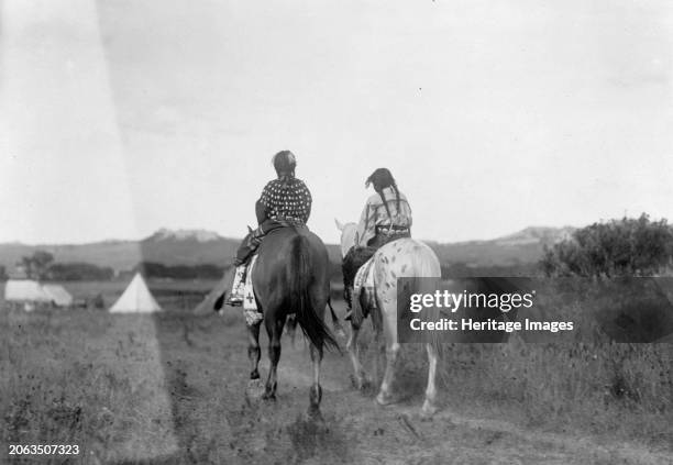 Two daughters of a chief on horseback, riding away from camera toward tents in background, circa 1907. Creator: Edward Sheriff Curtis.