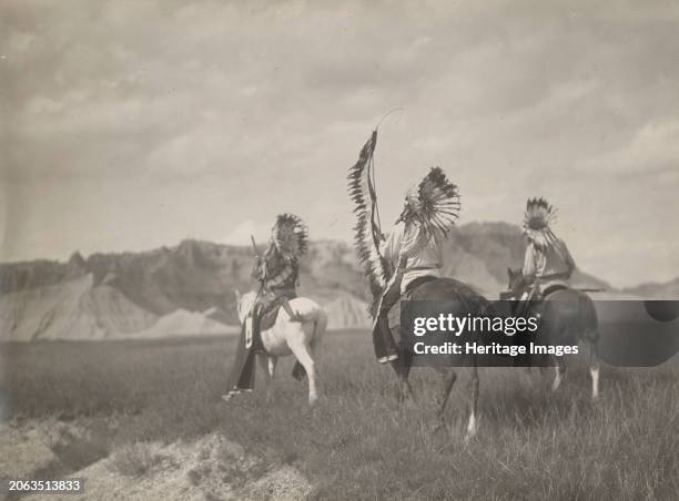 Sioux war party, 1905. Photograph shows three warriors, wearing headdress, riding on horseback, one holding a feathered lance and the others holding...