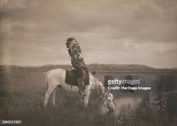An oasis in the Badlands, circa 1905. Photograph shows Red Hawk, an Oglala warrior, sitting on a horse that is drinking from a small pond in the...