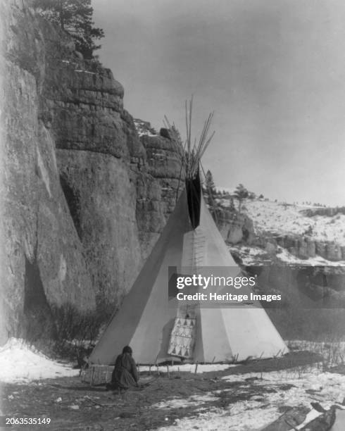 Hide stretching-Apsaroke, circa 1908. Apsaroke woman stretching hide that has been secured to the ground by stakes, tipi in background, cliff on...