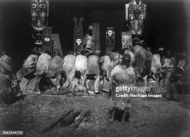 An incident of the Winter Dance-Nakoaktok, circa 1914. Kwakiutl people, some bowing before totem poles in background, others seated facing front as...