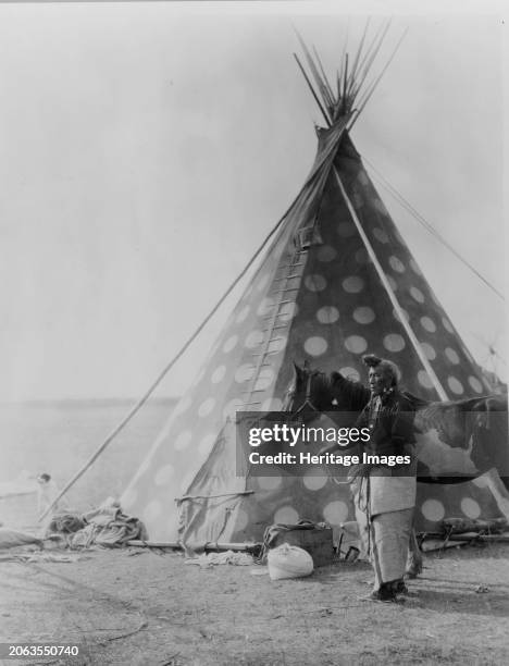 Blackfoot tepee, circa 1927. Blackfoot Indian, holding horse outside tipi. Creator: Edward Sheriff Curtis.