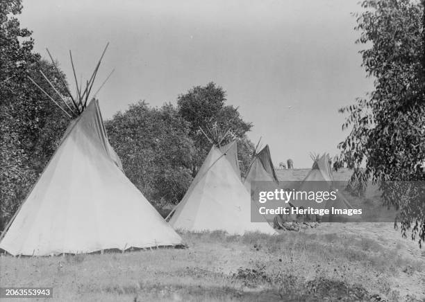 Camp life, circa 1908. Four canvas covered tipis, Assiniboine women and children seated on ground, South Dakota. Creator: Edward Sheriff Curtis.