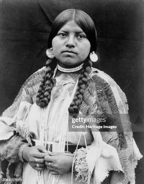 Cayuse woman, half-length portrait, standing, facing front, braids, shell disk earrings, shell bead choker and shell beads around neck, beaded...