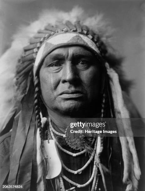 White Man Runs Him, Apsaroke, circa 1908. Portrait, man, head-and-shoulders, facing front, feather headress, shell concho and shell beads around...