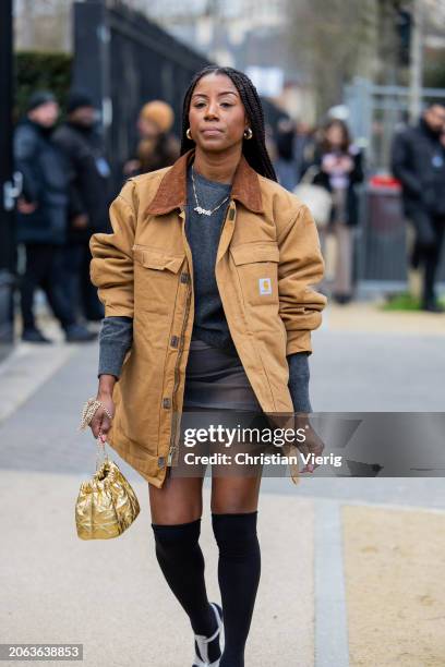 Guest wears beige oversized Carhartt jacket, grey jumper, skirt, golden bag, over knee high socks outside Lacoste during the Womenswear Fall/Winter...