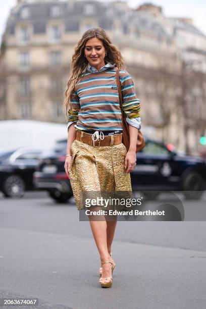 Emili Sindlev wears a brown and green striped polo shirt, a large brown leather belt, a leather gathered bag, a golden texture pleated knee length...