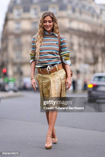 Emili Sindlev wears a brown and green striped polo shirt, a large brown leather belt, a leather gathered bag, a golden texture pleated knee length...