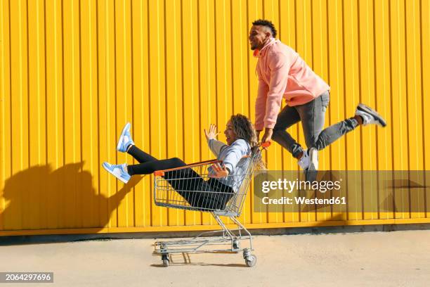 carefree woman sitting in shopping cart near man jumping in front of yellow striped wall - shopping cart stock pictures, royalty-free photos & images