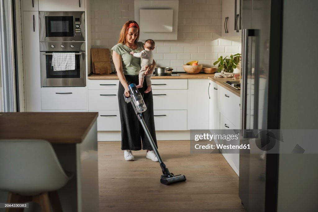 Woman carrying baby daughter and cleaning kitchen with vacuum cleaner