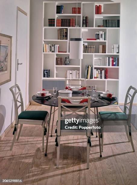 Interior view of a contemporary dining room, it contains a round table set for four, four wooden chairs with seat cushions and a pendant light...