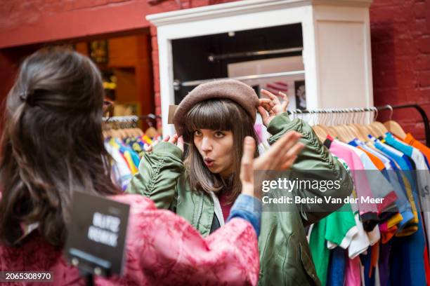 woman tries on hat in shop, while friend looks on. - feira da ladra mercado imagens e fotografias de stock