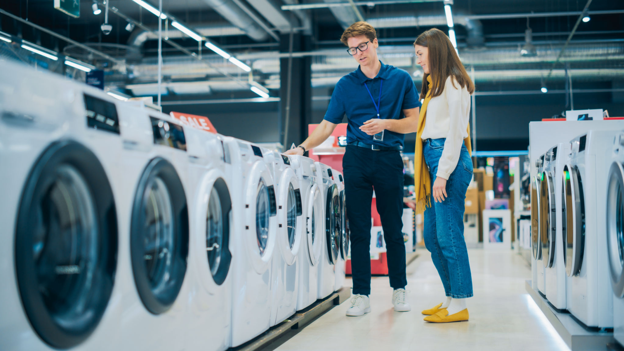 Portrait of a Female Customer Seeking Expert Advice from Retail Home Electronics Expert for Washing Machine Purchase. Young Woman Explores Contemporary Laundry Appliance Options in a Department Store Portrait of a Female Customer Seeking Expert Advice from Retail Home Electronics Expert for Washing Machine Purchase. Young Woman Explores Contemporary Laundry Appliance Options in a Department Store